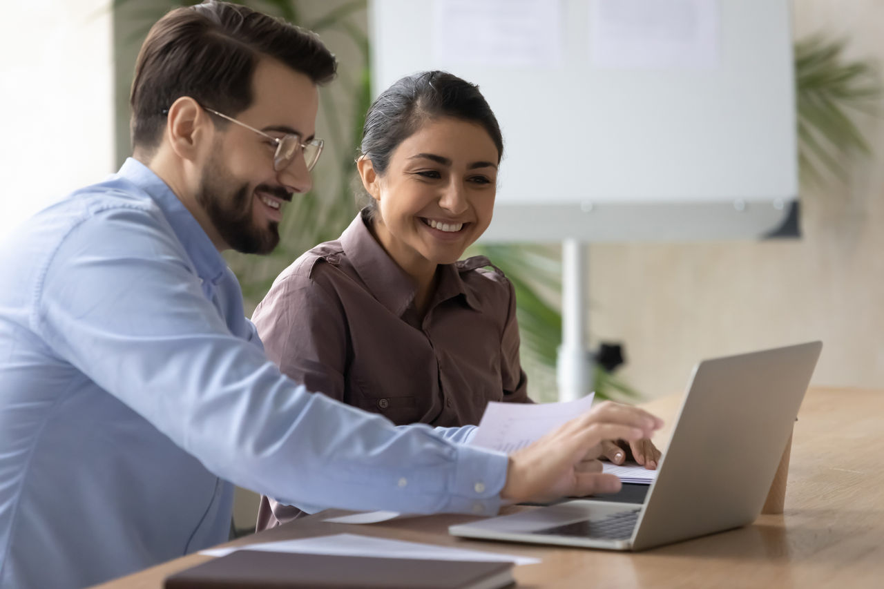 Smiling diverse colleagues working on project together, happy Caucasian businessman and Indian businesswoman sitting at desk, looking at laptop screen, discussing strategy, sharing ideas